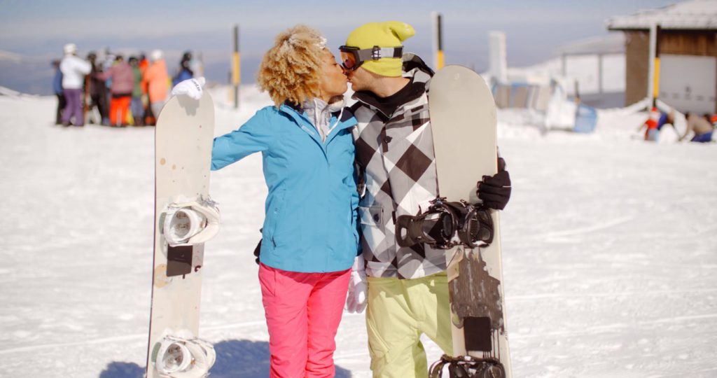 couple kissing on the ski slopes while holding snowboards